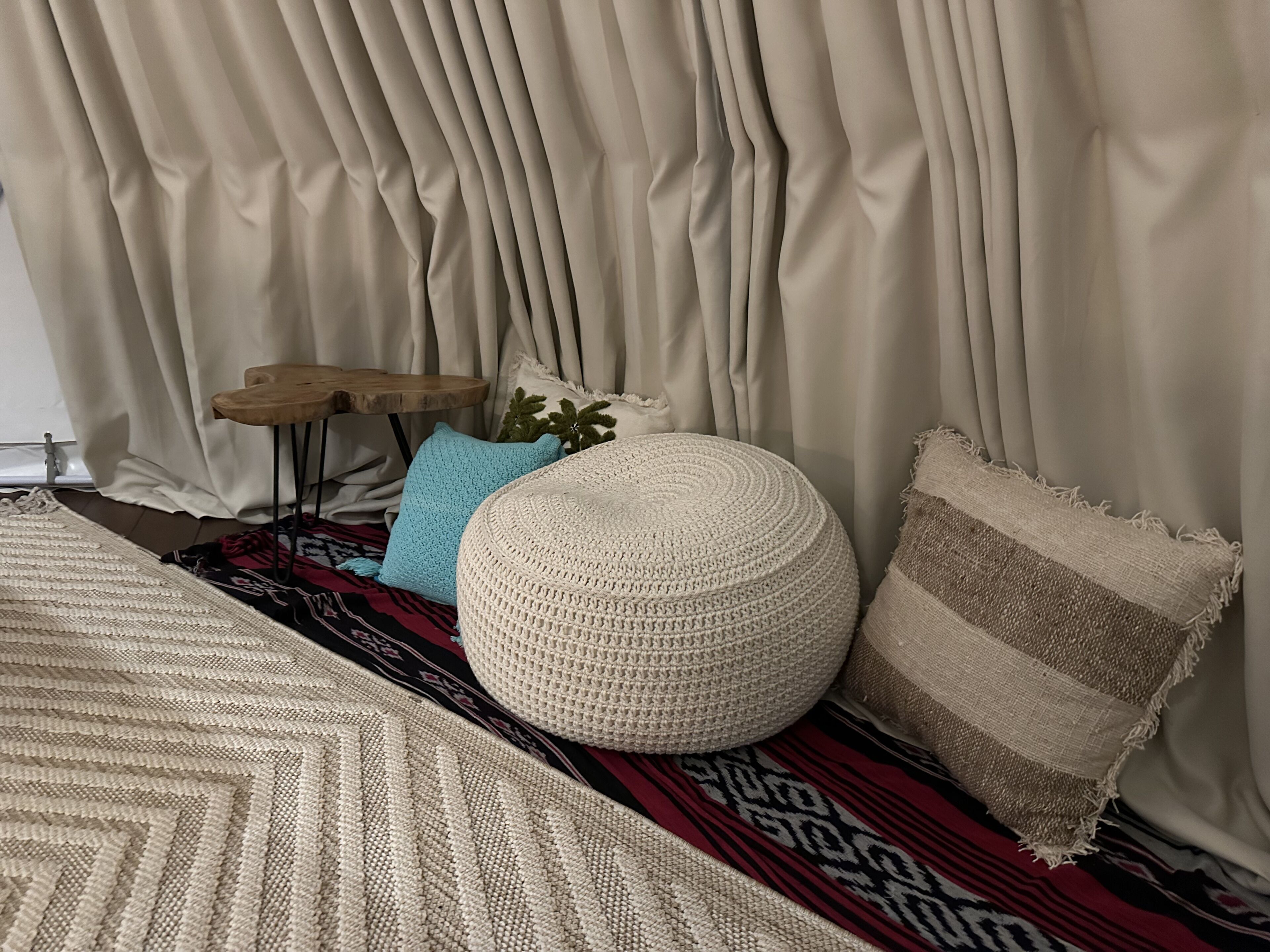 A room with a large beige curtain, a round beige ottoman, a blue pillow, and a patterned rug.