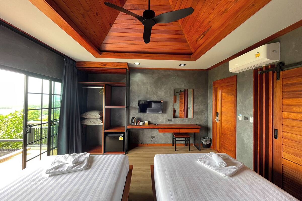 A hotel room with two beds, a desk, a ceiling fan, and a large window.