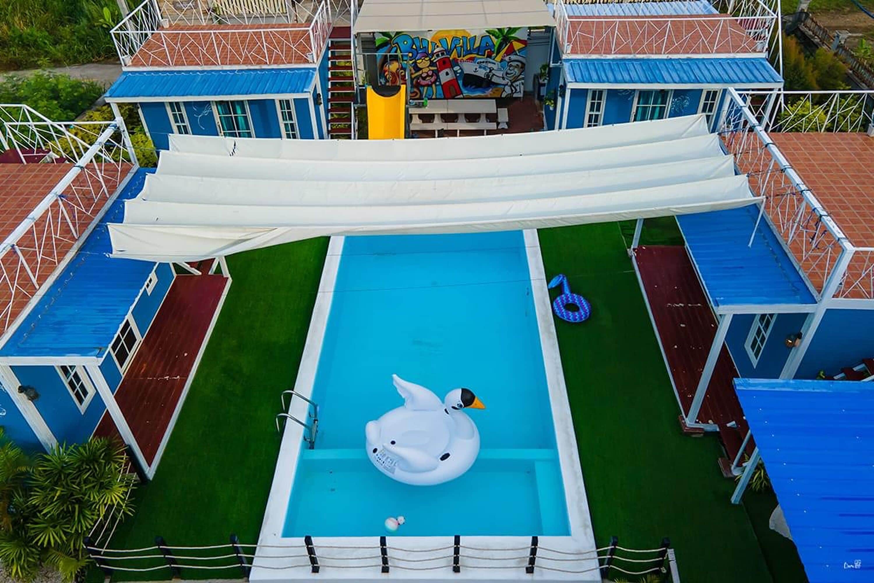 Una piscina con un flotador con forma de cisne blanco, rodeada de edificios azules y rojos con barandillas blancas.