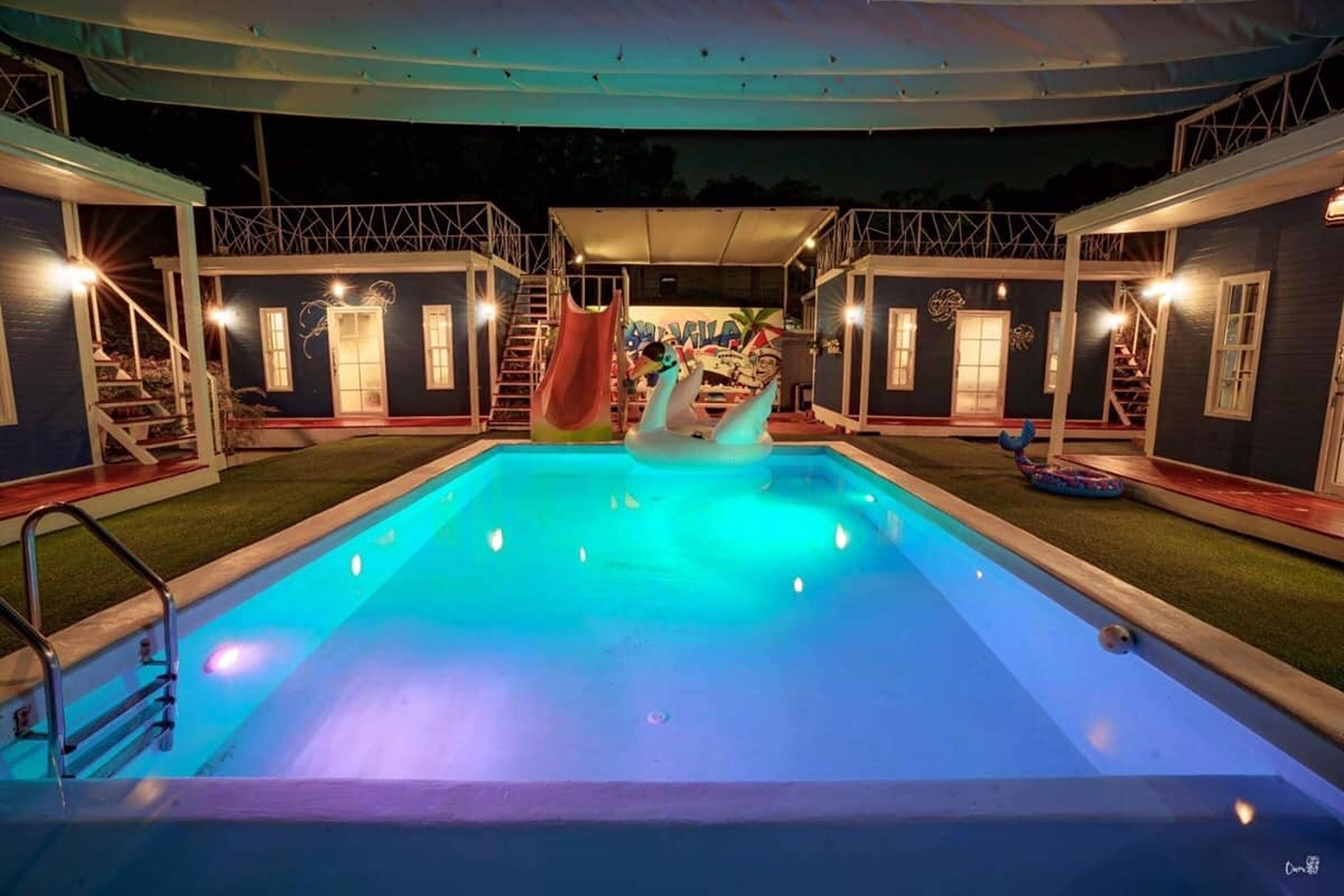 A swimming pool with a water slide, surrounded by two-story buildings with balconies and stairs.