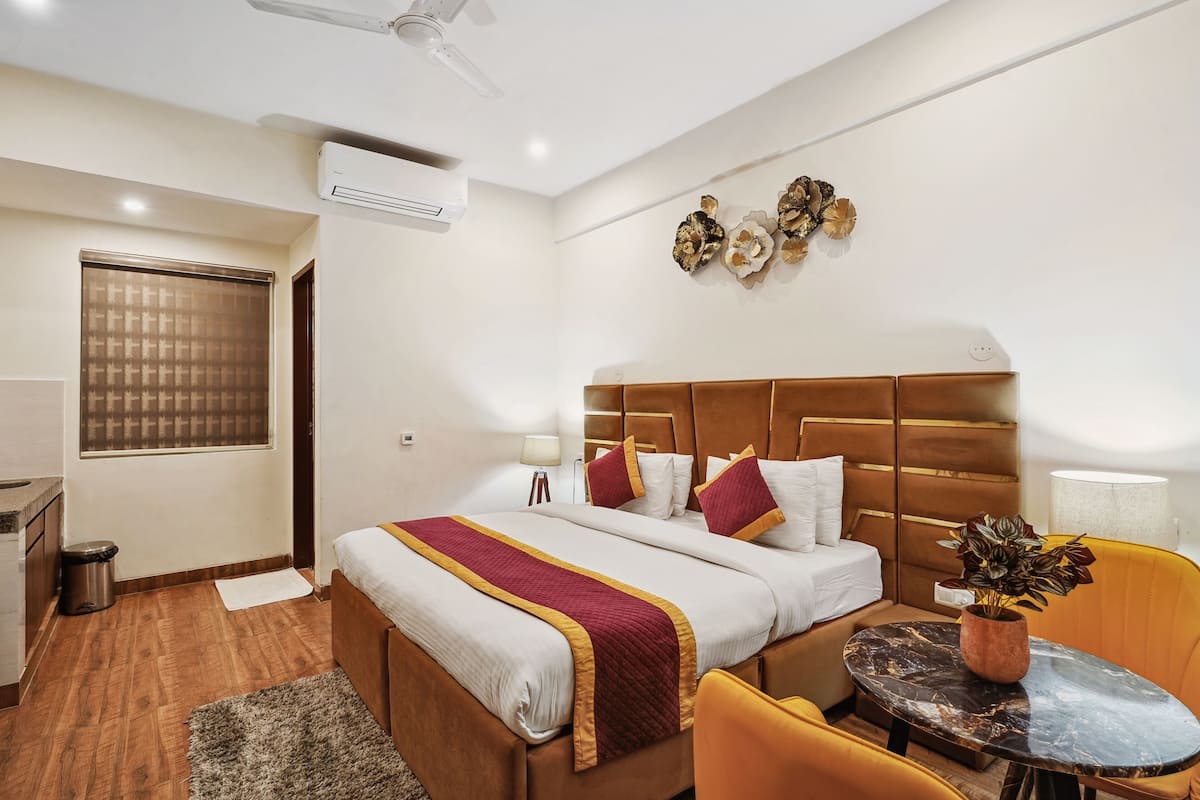 A hotel room with a large bed, a bedside table with a lamp, a small round table with a plant, and a window with blinds.