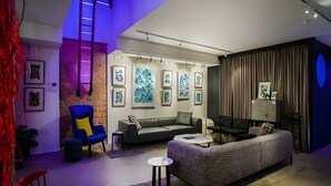 Una sala moderna con un sofá gris, un sillón azul, una mesita redonda y una mesa de centro negra. Hay cuadros enmarcados en la pared y un reloj también en la pared.