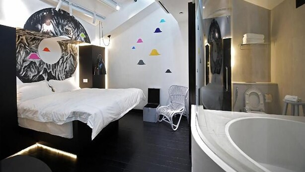 Una habitación de hotel moderna con una cama, una silla blanca, una bañera y una pared con formas geométricas coloridas.