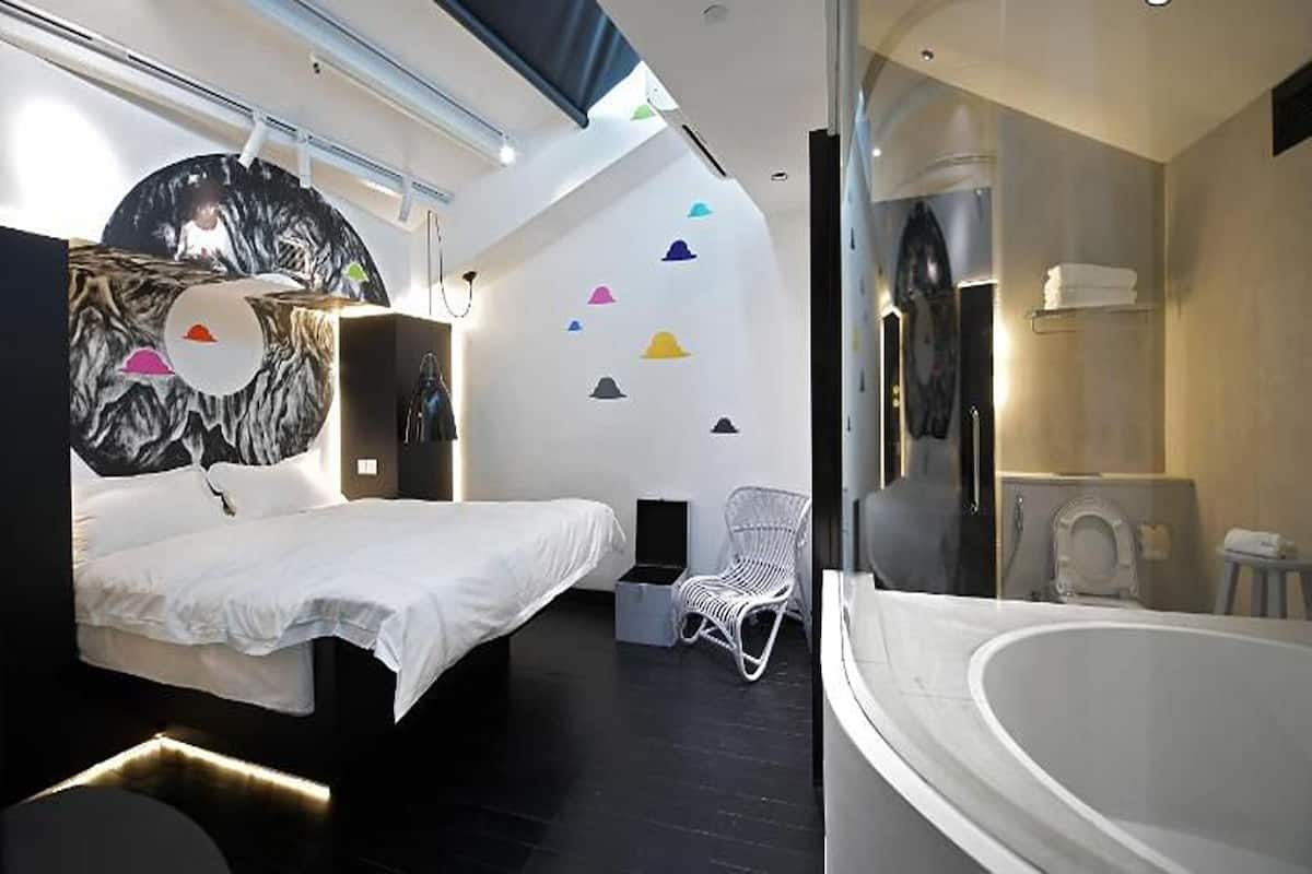 Une chambre d’hôtel moderne avec un lit, une chaise blanche, une baignoire et un mur orné de motifs géométriques colorés.