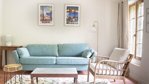 A living room with a light blue sofa, a wicker chair, a small table, and two framed posters on the wall. - Gîte Canterane, Petite Maison Provençale Rattachèe à Maison Sumiane, 4 Couchages (Brignoles)
