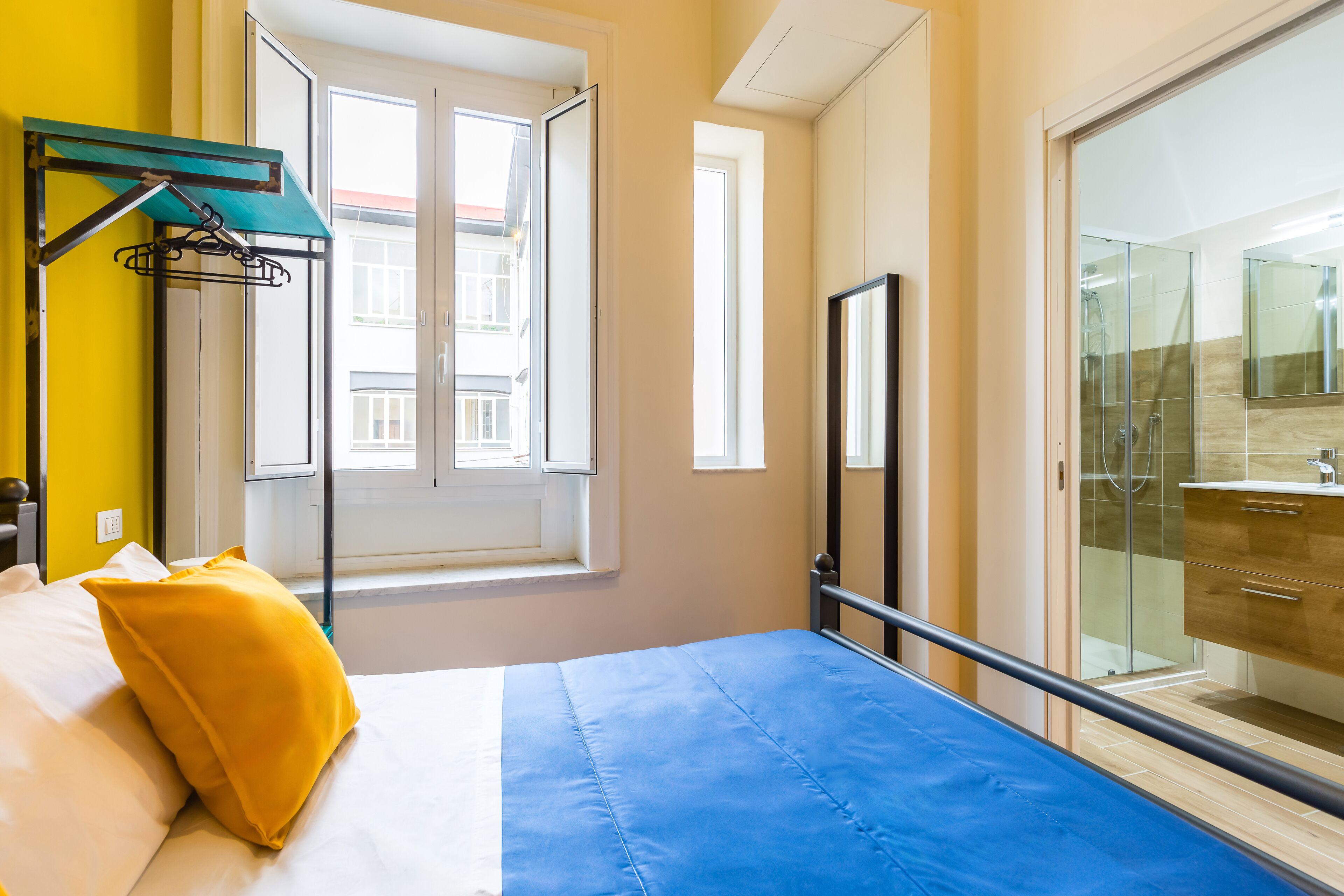 A bed with a blue mattress and a yellow pillow, a window with a view of buildings, a bathroom with a glass shower, and a door leading to a balcony.