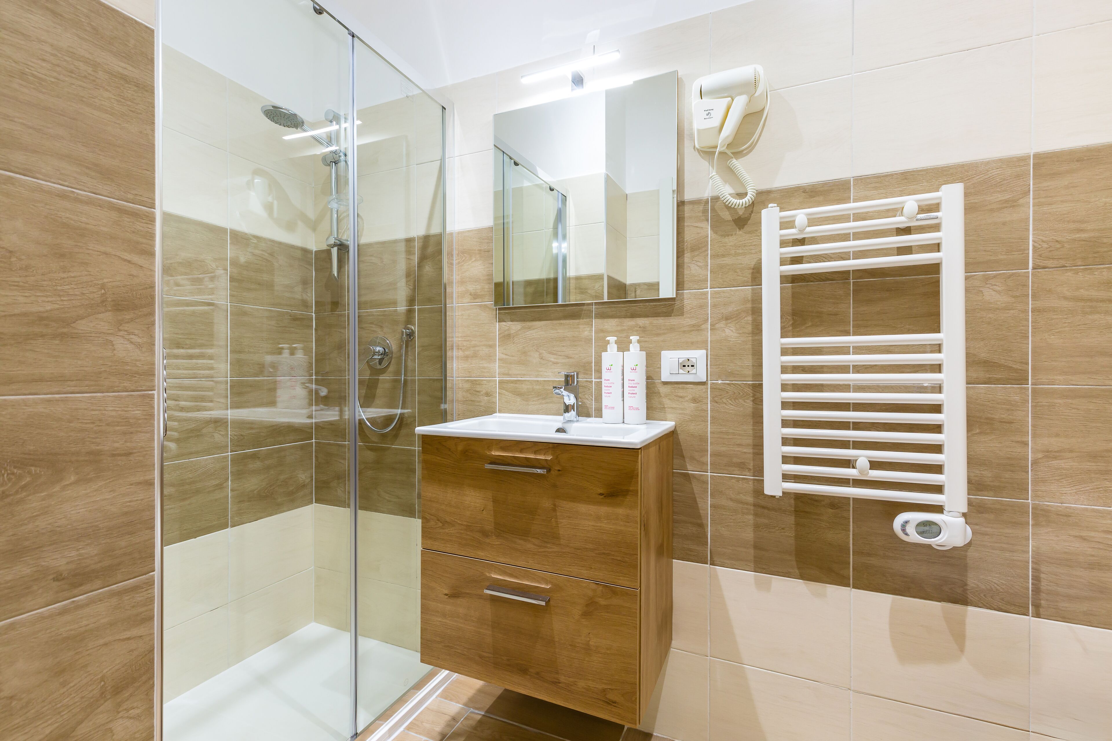 Ein Badezimmer mit einer gläsernen Duschkabine, einem Holzkabinet mit Schubladen, einem weißen Waschbecken und einem Handtuchhalter.