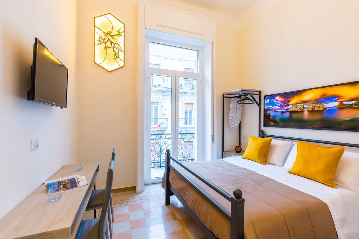 Quarto de hotel com cama, escrivaninha, televisão e varanda com vista para edifícios.