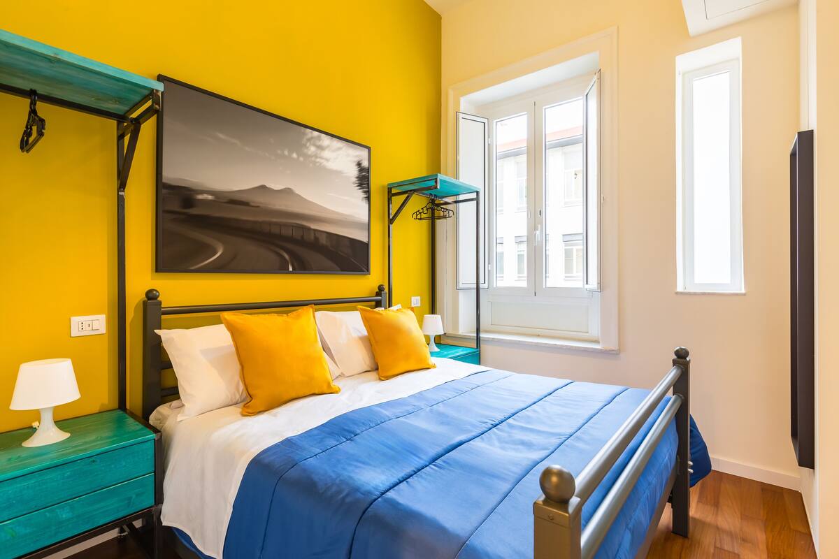 Quarto com cama, mesa de cabeceira, prateleira na parede, janela e porta.