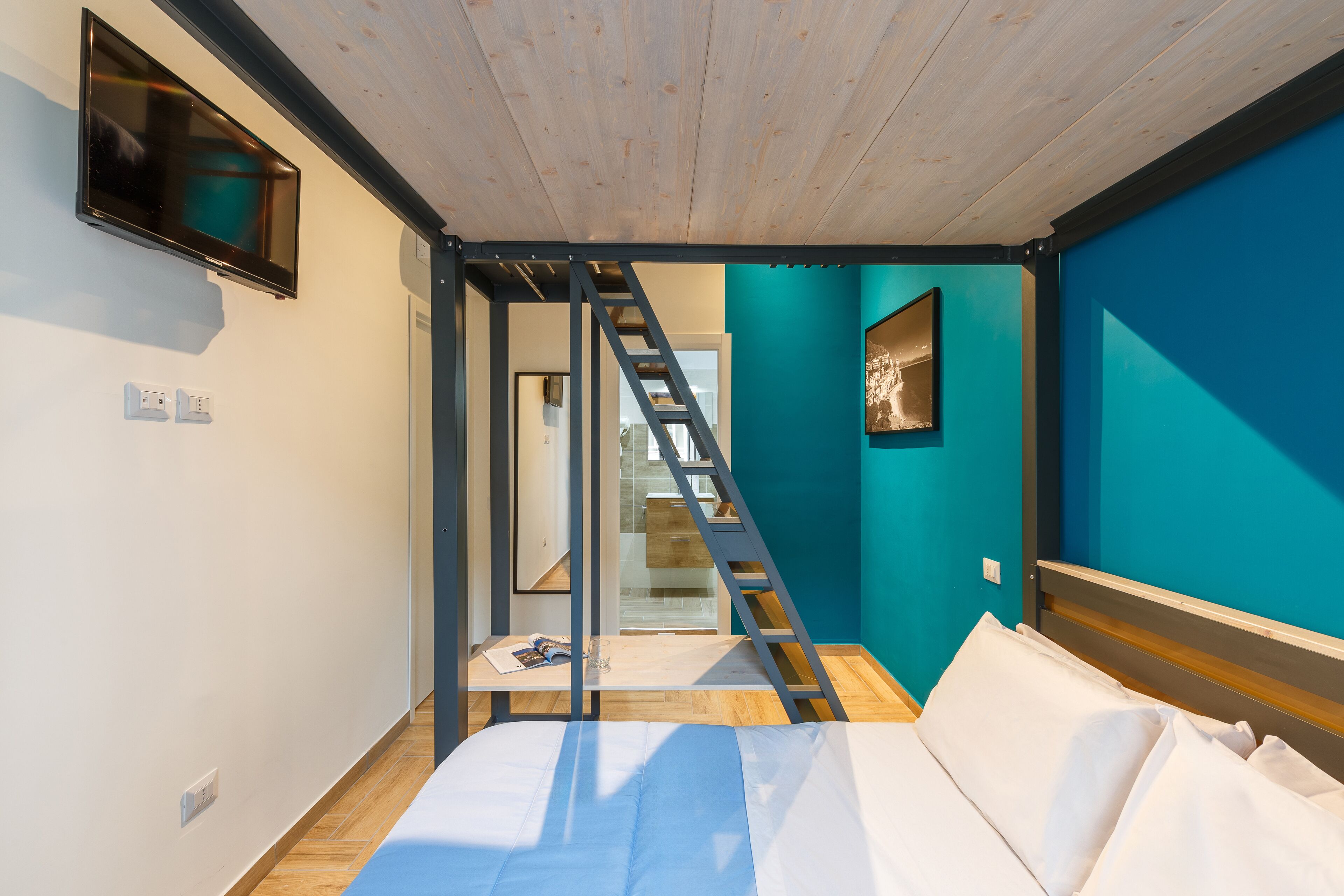 A bedroom with a bed, a TV mounted on the wall, a ladder leading to a staircase, and a blue accent wall.