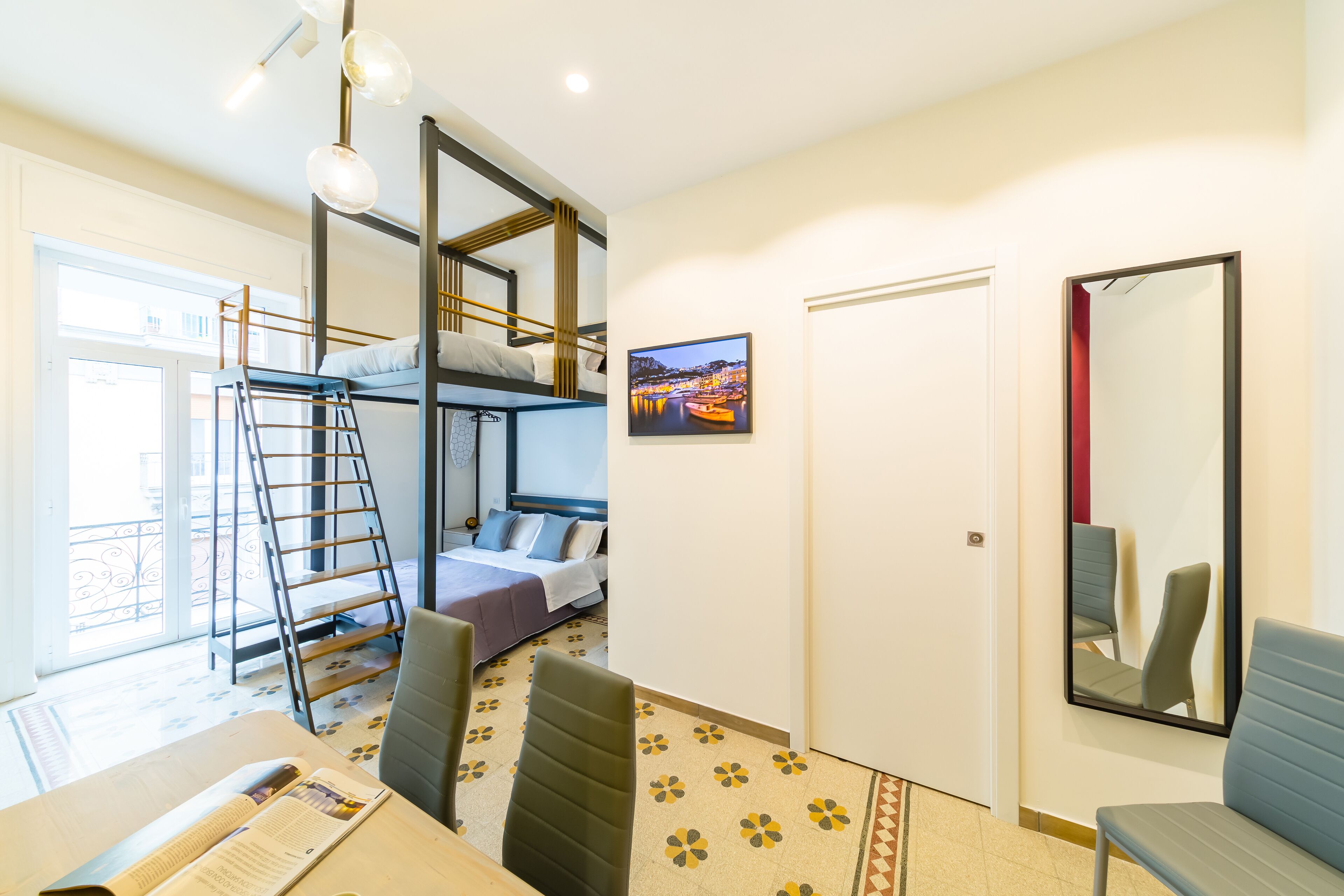 A room with bunk beds, a desk with a chair, a ladder, a door, a mirror, and a window with a view of a cityscape.