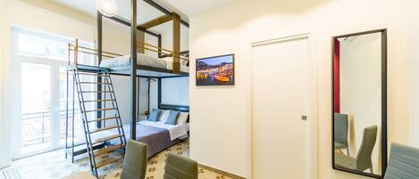 A room with bunk beds, a desk with a chair, a ladder, a door, a mirror, and a window with a view of a cityscape.