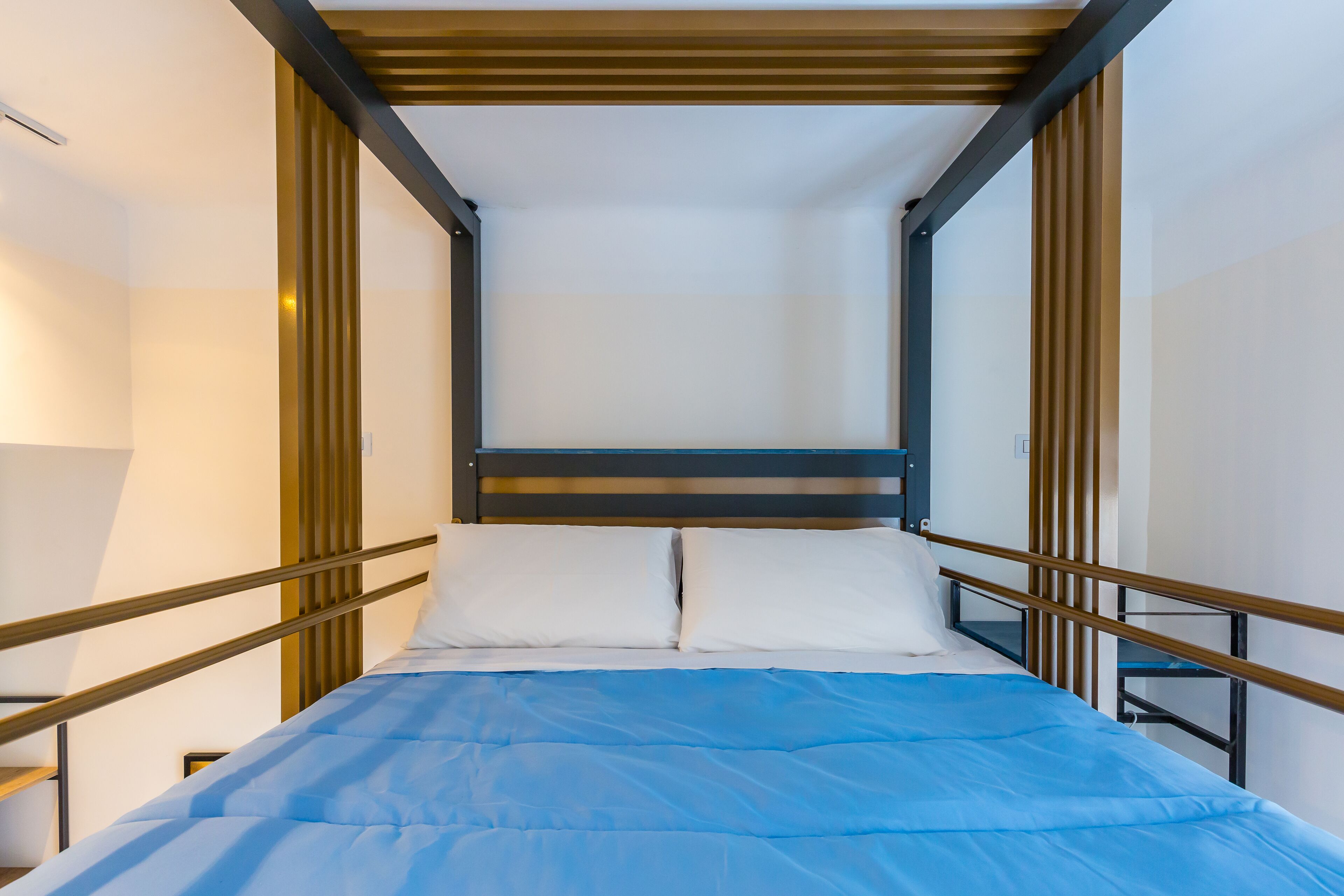 A modern, compact bed with a blue mattress and white pillows, framed by a wooden headboard and metal railings, in a minimalist room with white walls and a sloped ceiling.
