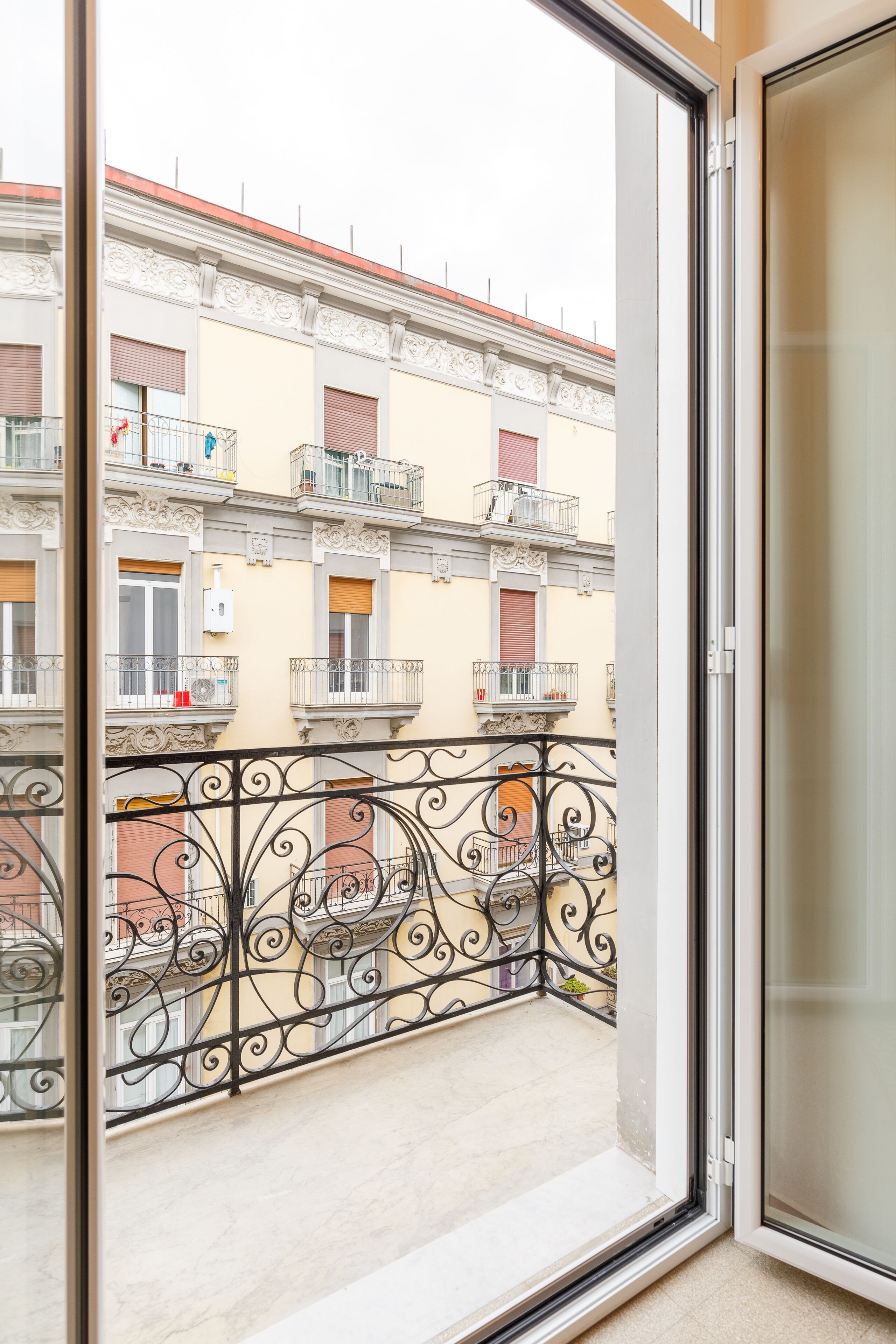 A balcony with ornate black wrought iron railings, a view of neighboring buildings with balconies, and a door leading to an interior space.