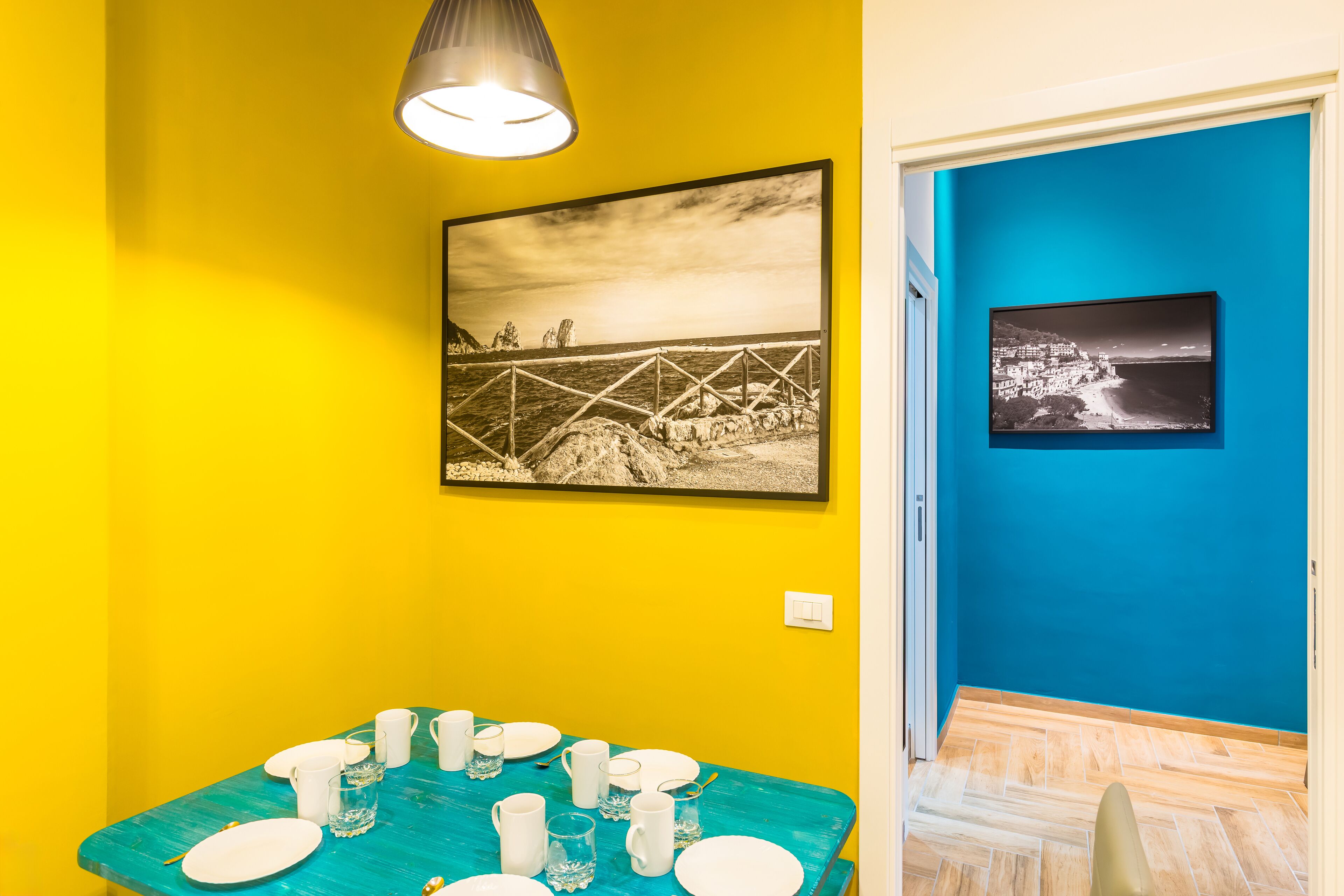 A dining area with a turquoise table set for six, two black and white photographs on the walls, and a bright yellow wall.