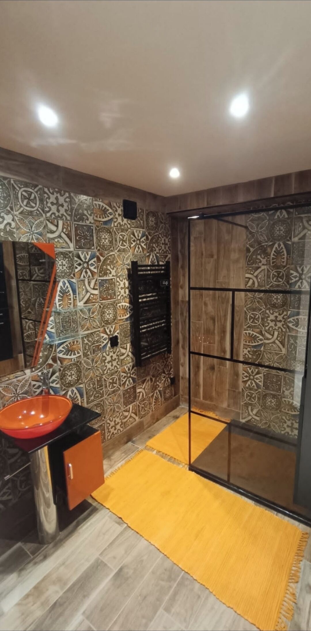A bathroom with patterned tiles, a glass shower enclosure, a sink with a red basin, and a yellow rug.