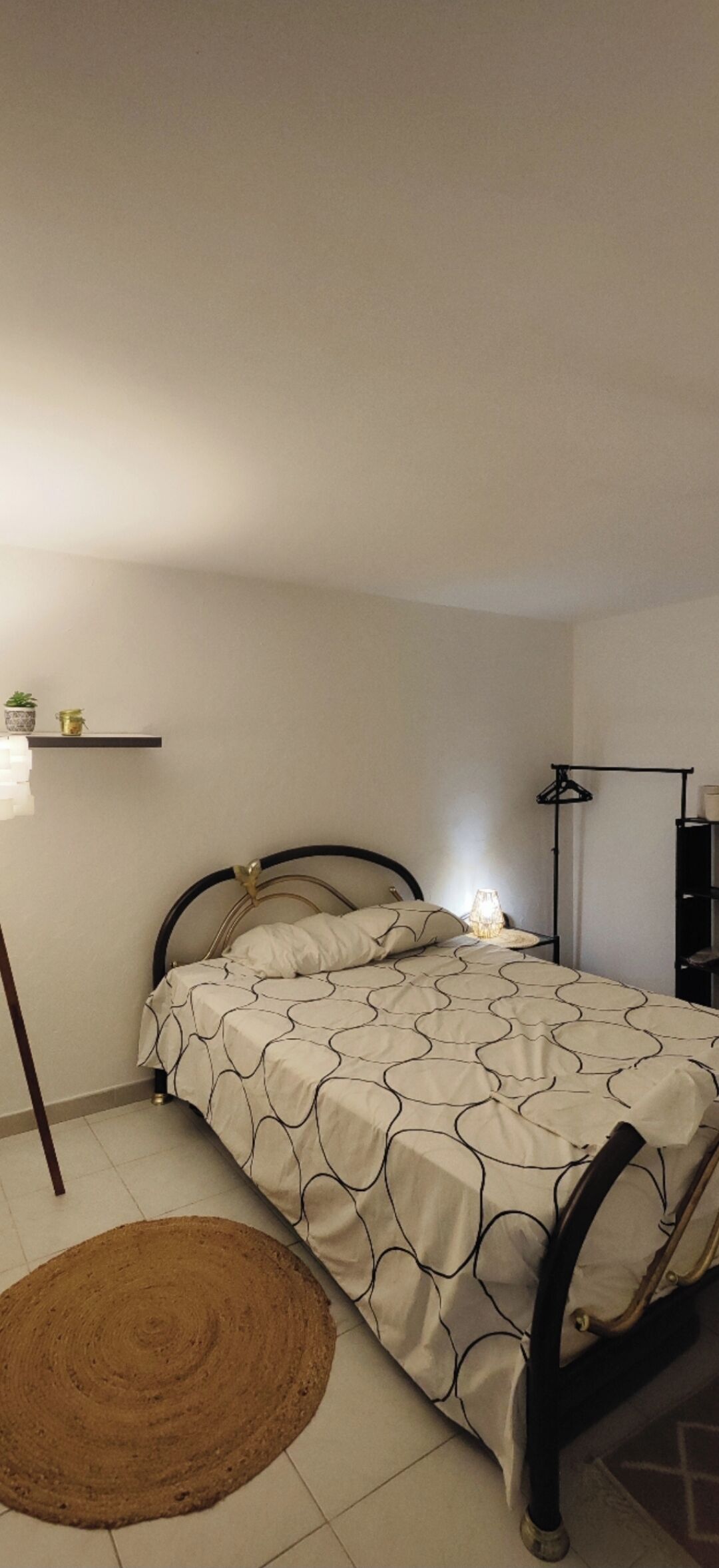 A small, neatly arranged bedroom with a single bed, a bedside table, a lamp, and a shelf with decorative items.
