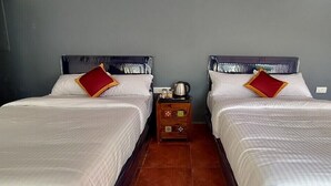 Habitación de hotel con dos camas, cada una con ropa de cama blanca y almohadas rojas, una mesita de noche de madera con una tetera, y suelo de baldosas.