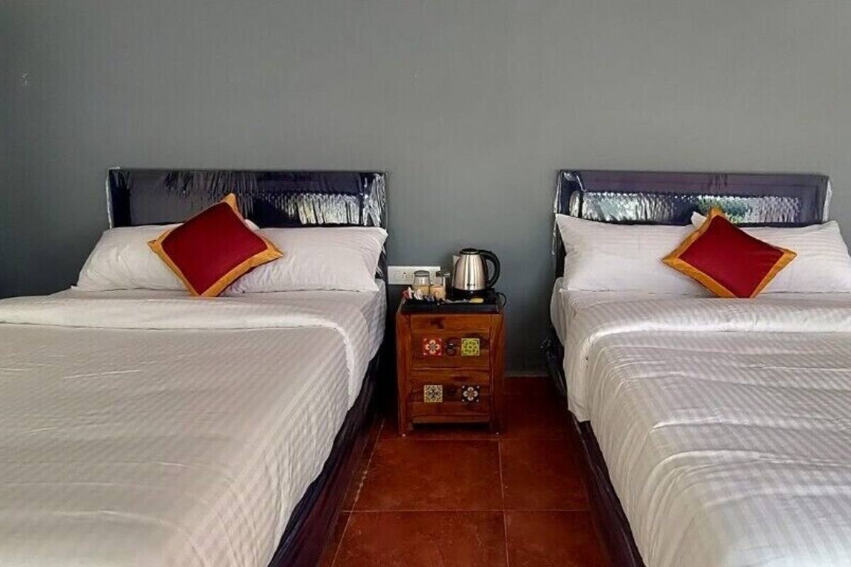 A hotel room with two beds, each with white bedding and red pillows, a small wooden nightstand with a kettle, and a tiled floor.
