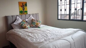 Una cama bien tendida con ropa blanca y dos almohadas estampadas, un cabecero tapizado en gris y una ventana con rejas.