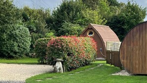 Une allée de gravier mène à un bâtiment en bois doté d’une fenêtre circulaire, entouré d’une pelouse bien entretenue et d’une haie ornée de fleurs rouges.