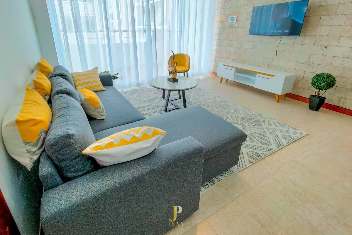 A modern living room with a grey sectional sofa, a small round table, and a TV mounted on a brick wall.