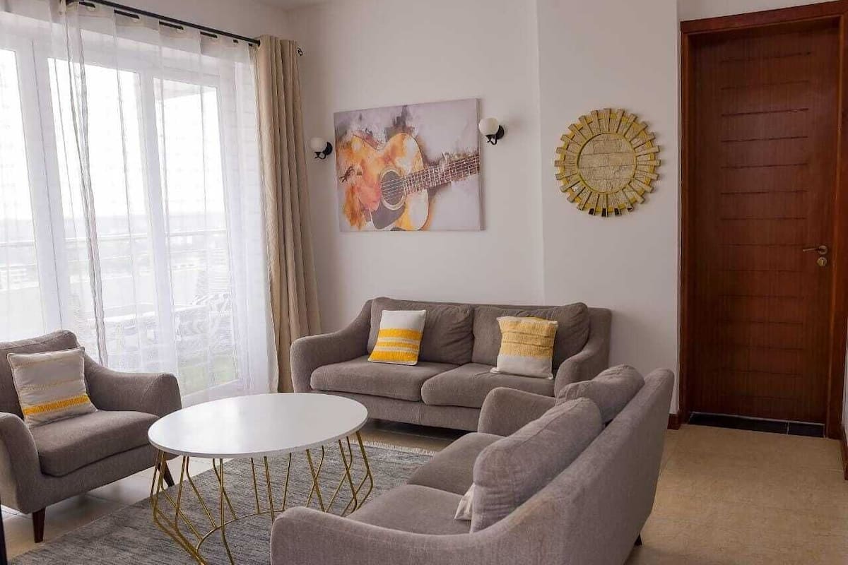 A living room with a grey sofa, two armchairs, a round coffee table, and a painting of a guitar on the wall.
