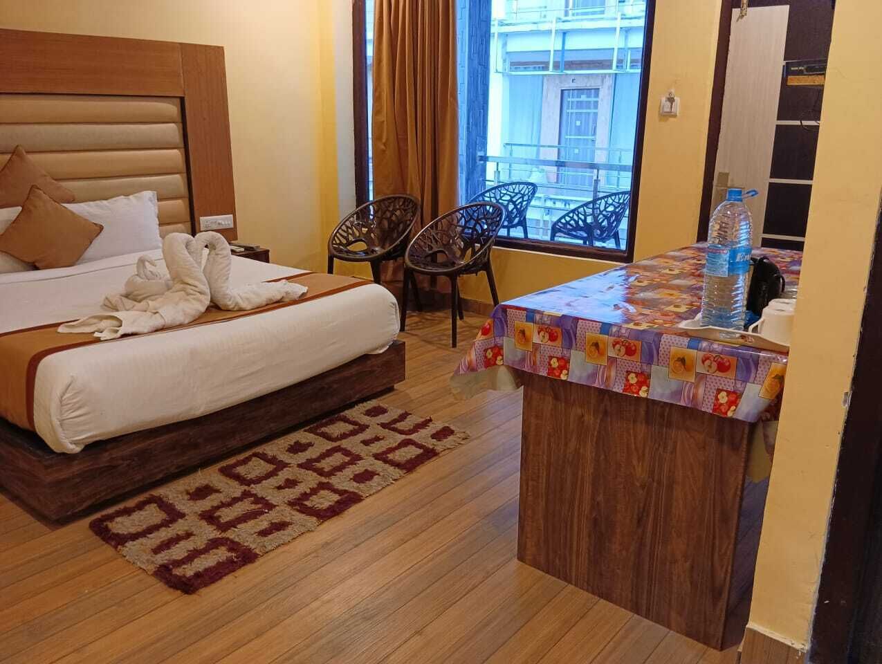 A hotel room with a bed, a small desk with a table, two chairs, a window with curtains, and a patterned rug.