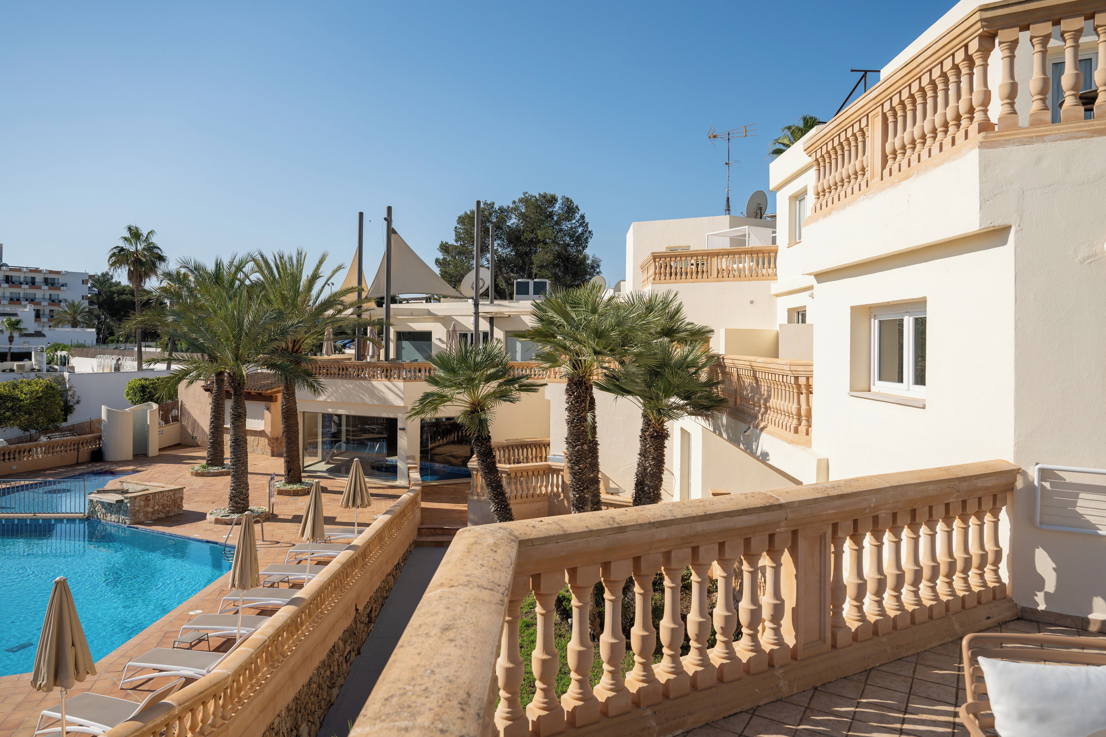 A pool area with lounge chairs and umbrellas, a building with balconies, and palm trees.