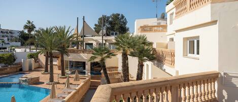 Un área de piscina con sillones de descanso y sombrillas, un edificio con balcones y palmeras.
