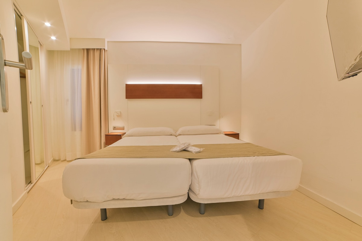 A hotel room with a double bed, a bedside table, a nightstand, and a wall-mounted light fixture.