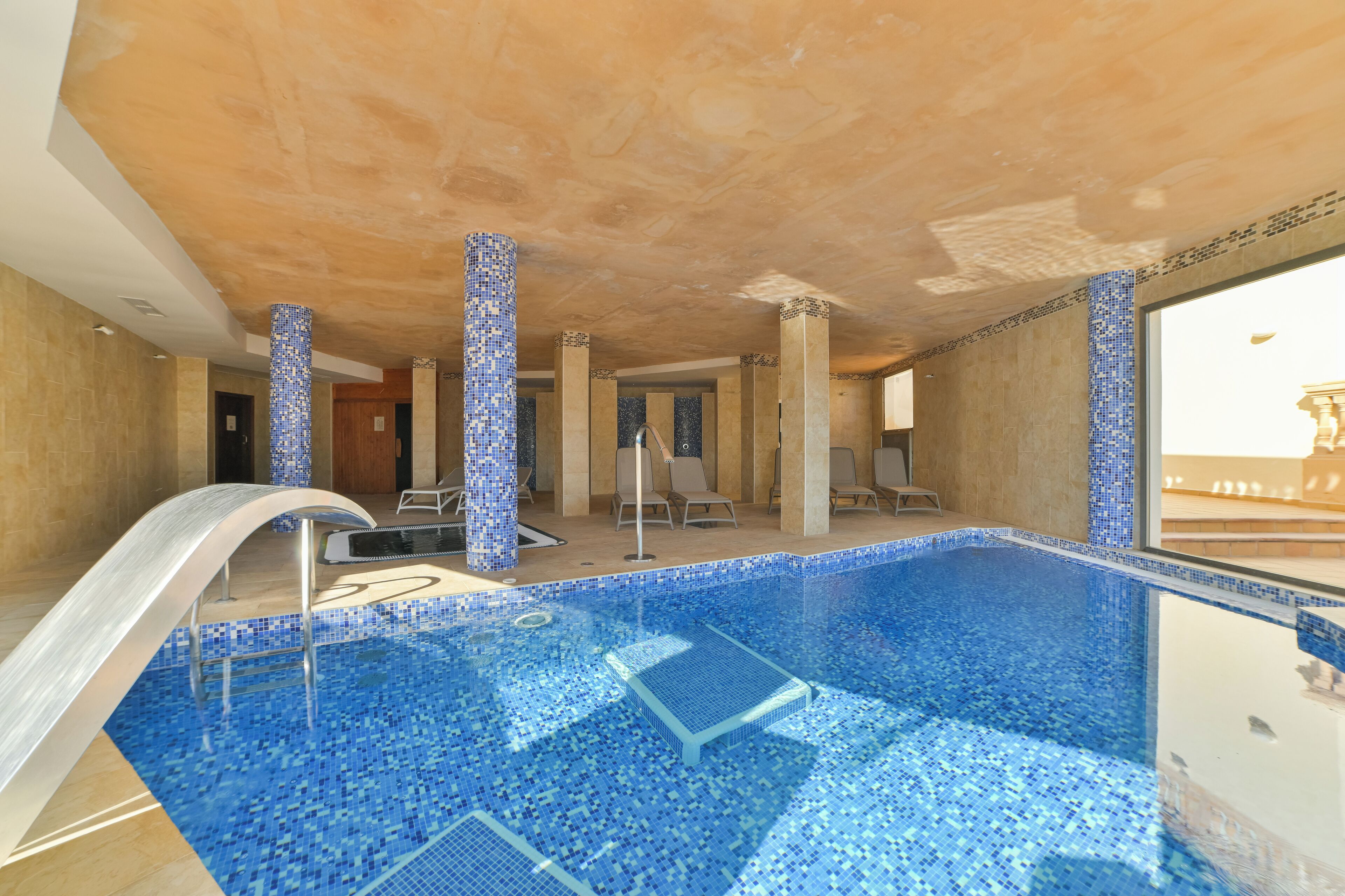 Une piscine intérieure aux carreaux de mosaïque bleus, dotée d’un plongeoir et de chaises longues.