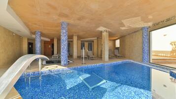 Une piscine intérieure aux carreaux de mosaïque bleus, dotée d’un plongeoir et de chaises longues.