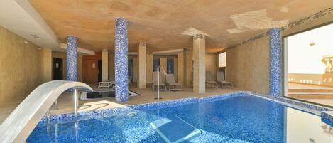 An indoor swimming pool with blue mosaic tiles, a diving board, and lounge chairs.