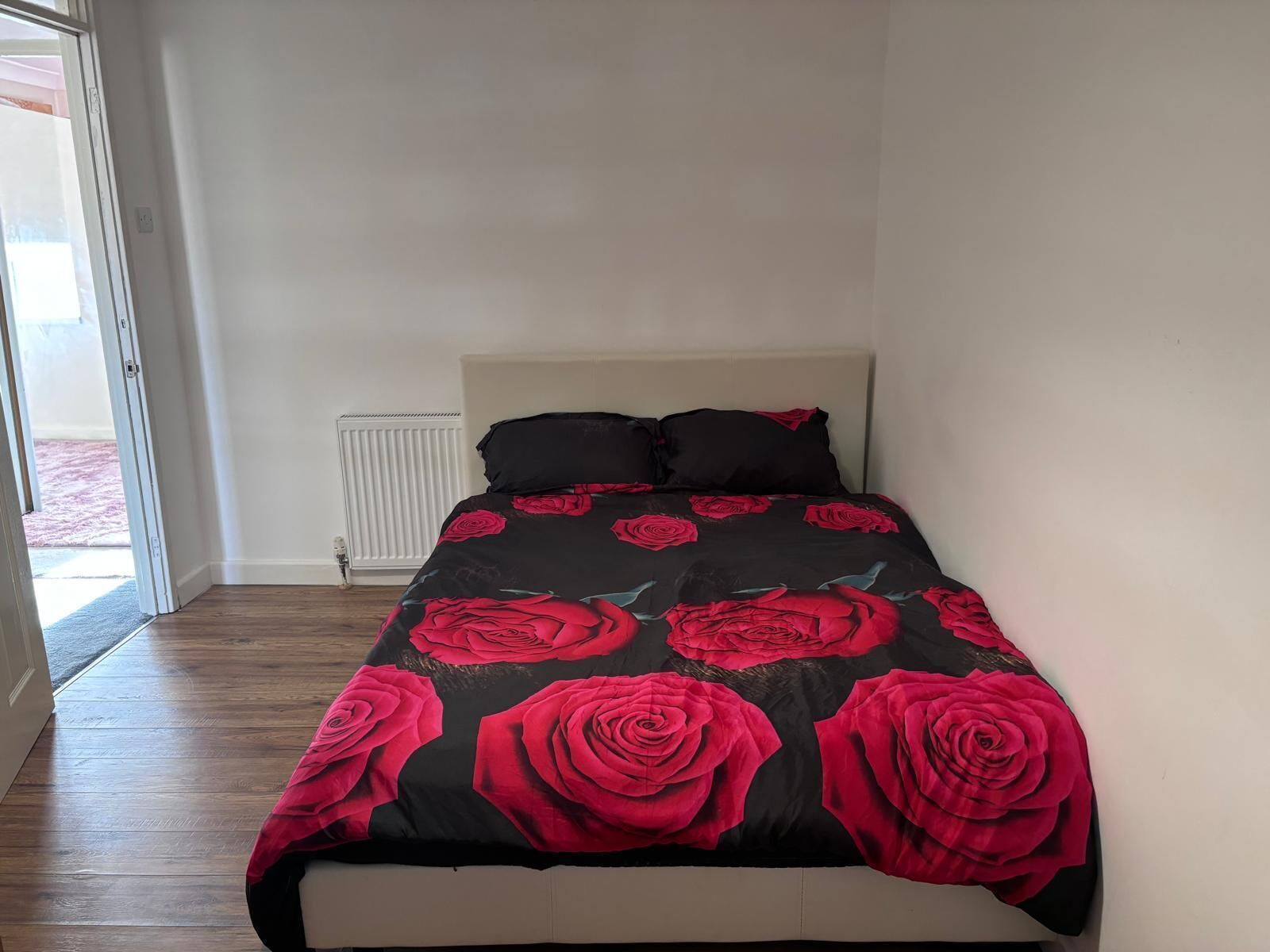 Une chambre à coucher avec un lit recouvert d’une courtepointe à motifs de roses rouges et noires, une tête de lit blanche et un radiateur fixé au mur.