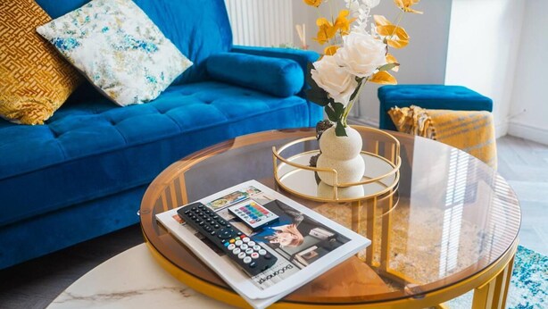 Ett vardagsrum med en blå soffa, ett soffbord i glas med en tidning och fjärrkontroller, och en vas med blommor.