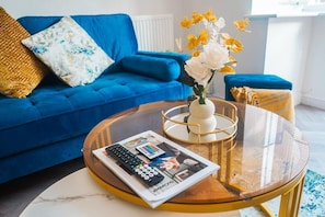 A living room with a blue sofa, a glass coffee table with a magazine and remote controls, and a vase with flowers. - Immaculate 4-bed House in Kings Norton (Birmingham)