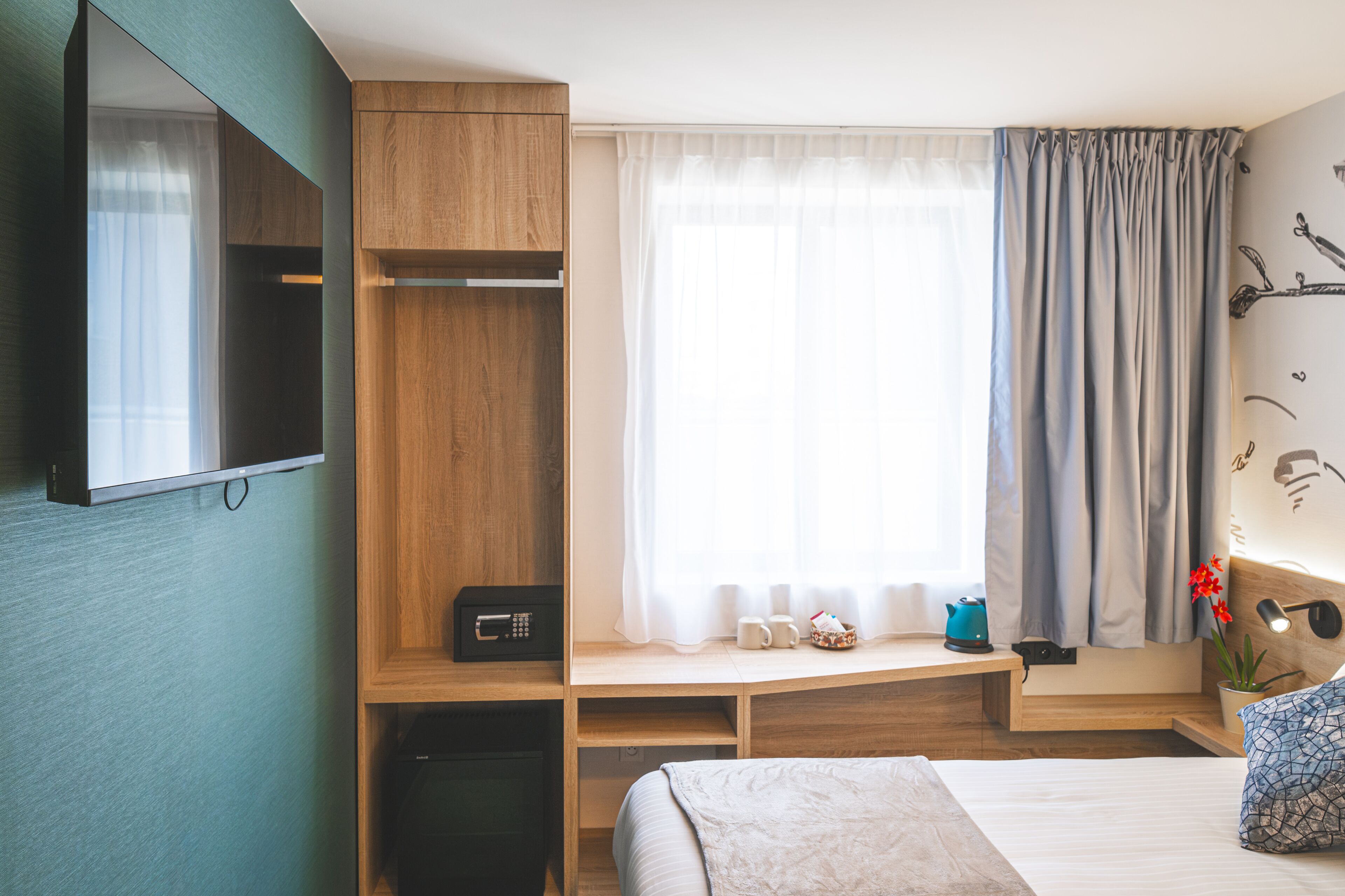 A hotel room with a bed, a wooden desk, a TV mounted on the wall, a window with curtains, and a small plant on the bedside table.