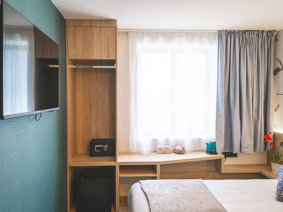 A hotel room with a bed, a wooden desk, a TV mounted on the wall, a window with curtains, and a small plant on the bedside table.