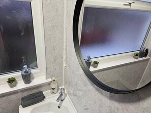 Ein Badezimmer mit einem weißen Waschbecken, einem Spiegel und einem Fenster mit Milchglas. Auf der Fensterbank stehen kleine Pflanzen und auf dem Waschbecken eine Lotionflasche.