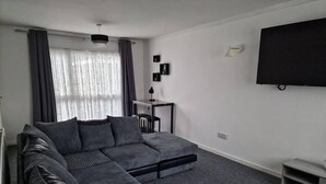 Un salon moderne avec un canapé d’angle, un téléviseur à écran plat fixé au mur et une petite table d’appoint.