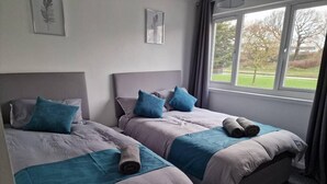 Une chambre avec deux lits, chacun doté d’une tête de lit, de draps gris et d’oreillers bleus. Des tableaux encadrés ornent le mur, et une fenêtre est agrémentée de rideaux.