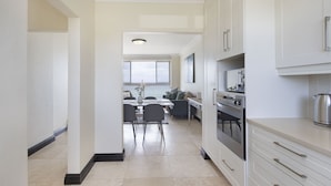 A modern kitchen with white cabinets, a stainless steel oven, and a dining area with a table and chairs. - Blouberg Heights 603 (Cape Town)