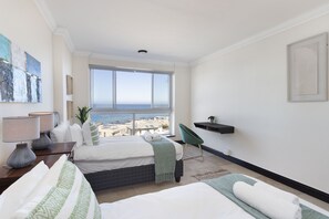 Une chambre d’hôtel avec deux lits, une vue sur la mer, un bureau avec une lampe et un fauteuil vert.