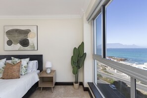 A bedroom with a bed, a bedside table, a lamp, a large potted plant, and a view of the ocean through a large window. - Blouberg Heights 603 (Cape Town)