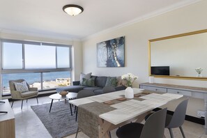A modern living room with a large window overlooking the sea, a grey sofa, a dining table, and a large mirror. - Blouberg Heights 603 (Cape Town)