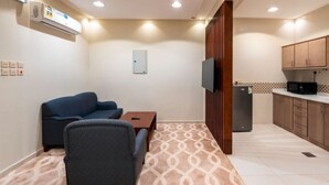 A compact living space with a sofa, armchair, small table, kitchenette, and a TV mounted on the wall. - Ninety Two (Unaizah)