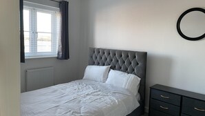 Une chambre à coucher comprenant un lit, une fenêtre avec des rideaux, un radiateur, une table de chevet avec des tiroirs et un miroir fixé au mur.