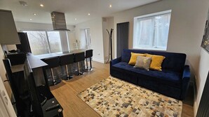 Una sala de estar moderna con un sofá azul oscuro, un tapete estampado y una zona de cocina con una mesa de comedor y taburetes de bar.