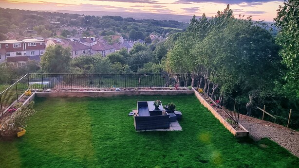 Un jardín bien cuidado con un banco de madera y una pequeña mesa, rodeado por una valla de metal.